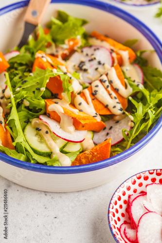 Bright vegan salad with arugula, sweet potato, radish, tahini dressing and cucumber, white background. Healthy vegan food concept.