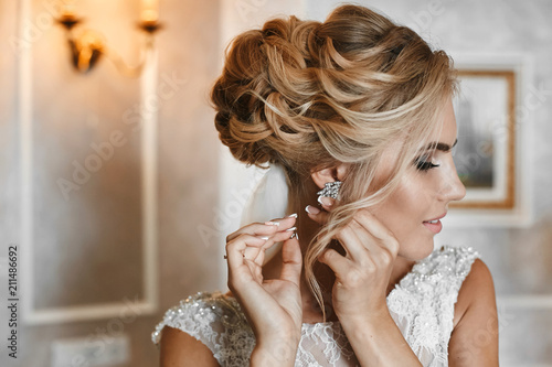 Beautiful, fashionable and elegant blond model girl with stylish wedding hairstyle, in lace white dress puts on her earring and posing at interior, wedding preparation of young bride