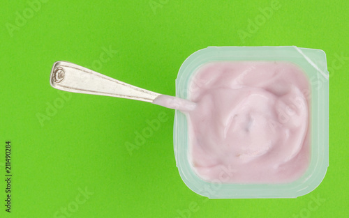 Yogurt in plastic cup close up with small silver spoon, top view photo of strawberry yoghurt on green background