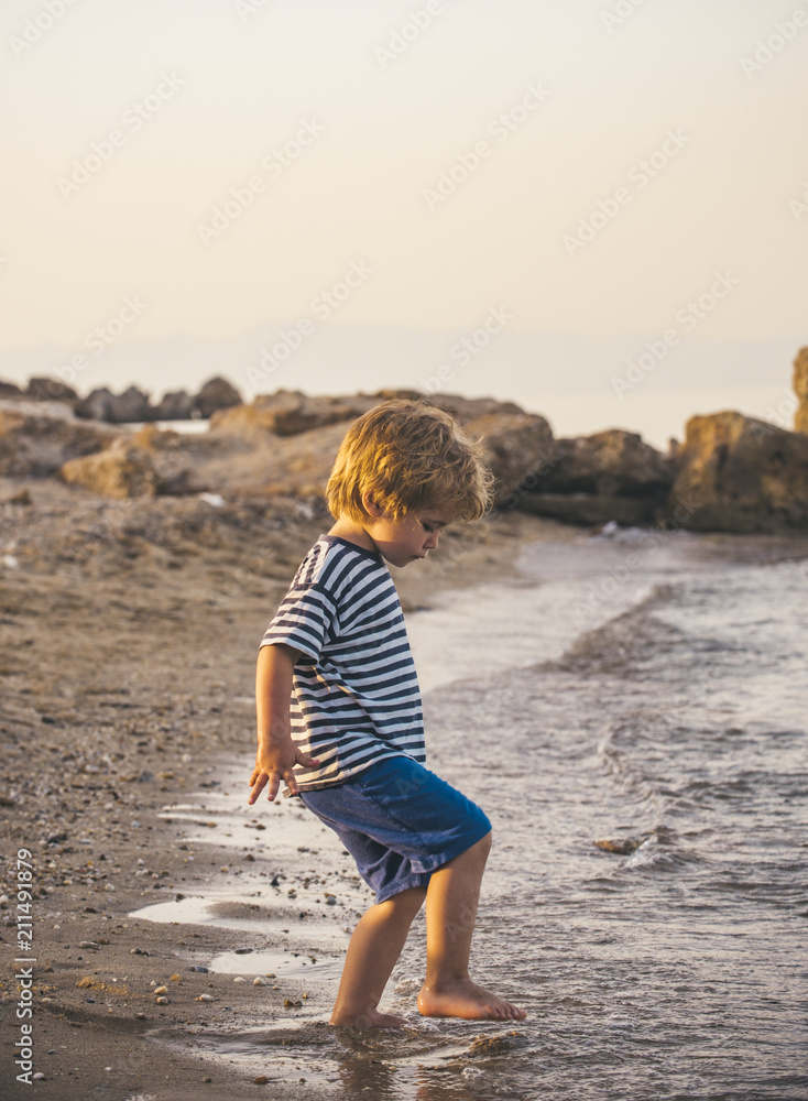 Child is walking by the sea, kid is playing with water on the beach. The tide in the ocean, a summer walk. Child and nature, sea shore, summer, tourism, vacation with child. Lonely boy on shoreline