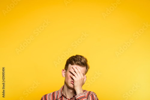facepalm. ashamed abashed man covering his face with hand. portrait of a young guy on yellow background popping up or peeking out from the bottom. copy space for advertising.