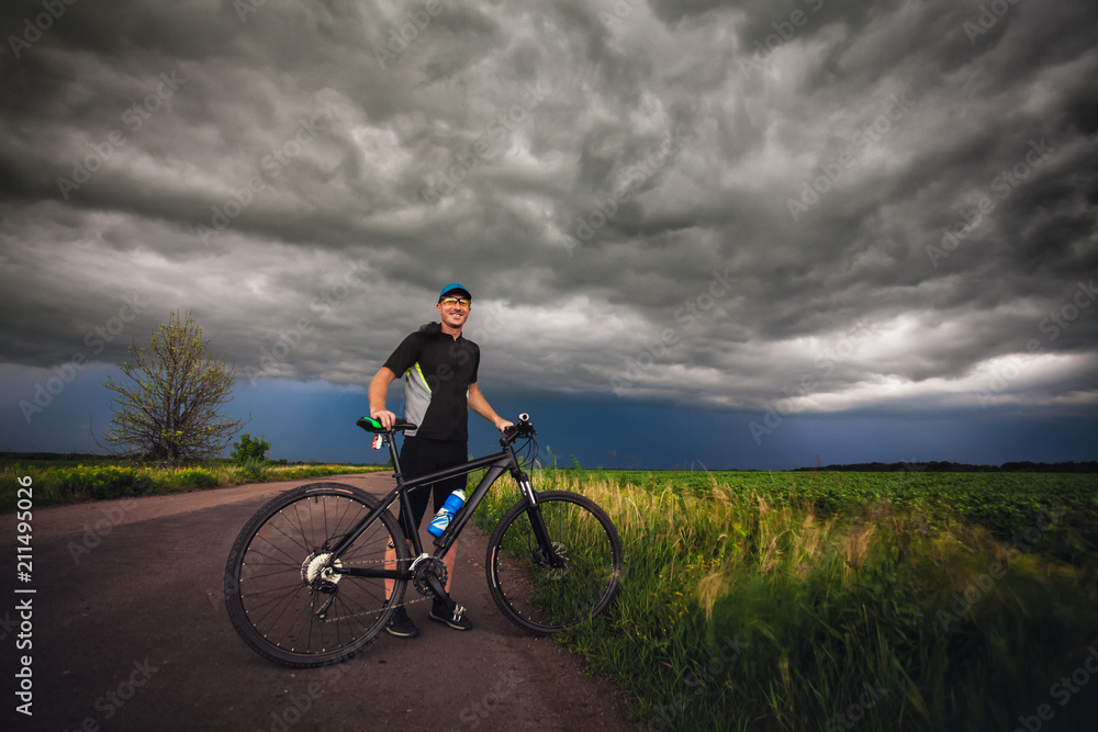 Fototapeta premium Happy man cyclist rides field road on a mountain bike. before the storm