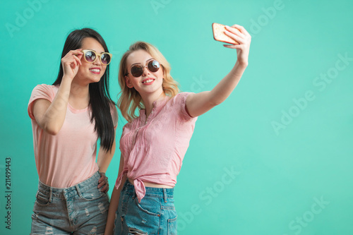 diversity, race, ethnicity, relationship concept - happy female couple, blonde caucasian and brunette Thai taking selfie with smartphone over blue background with copyspace