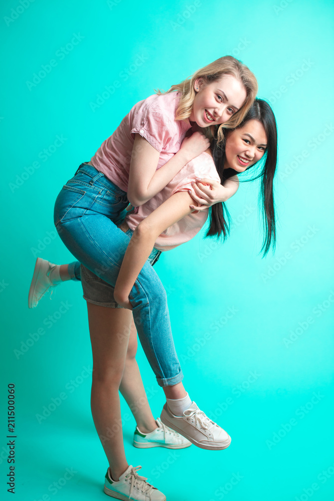 Two playful interacial girls piggybacking and having fun at studio blue background Two playful interacial girls piggybacking and having fun at studio blue background