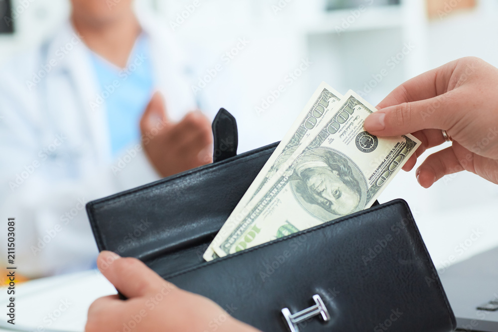 Fototapeta premium Female patient takes out of her wallet hundred dollar bills to pay for the services of a doctor. Bribery and corruption in Health Care Industry.