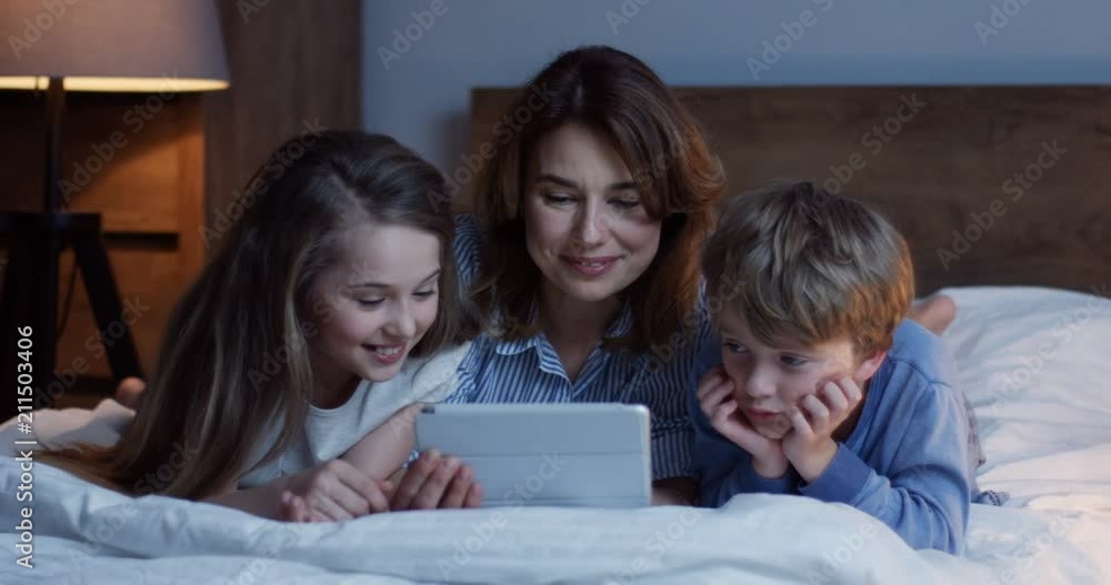 Portrait shot of the cheerful Caucasian mother and small children. boy and girl, lying on the unmade bed and laughing while watching funny video or cartoon on the tablet computer. Indoor. Portrait shot of the cheerful Caucasian mother and small children. boy and girl, lying on the unmade bed and laughing while watching funny video or cartoon on the tablet computer. Indoor.