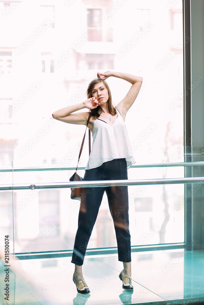 Stylish woman standing in shopping centre near huge window wearing white blouse and blue pants. Fashionable young model. Stylish woman standing in shopping centre near huge window wearing white blouse and blue pants. Fashionable young model.