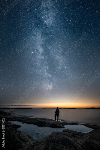 Silhouette of a man standing alone by a sea and looking at sunset and the stars of the epic milky way