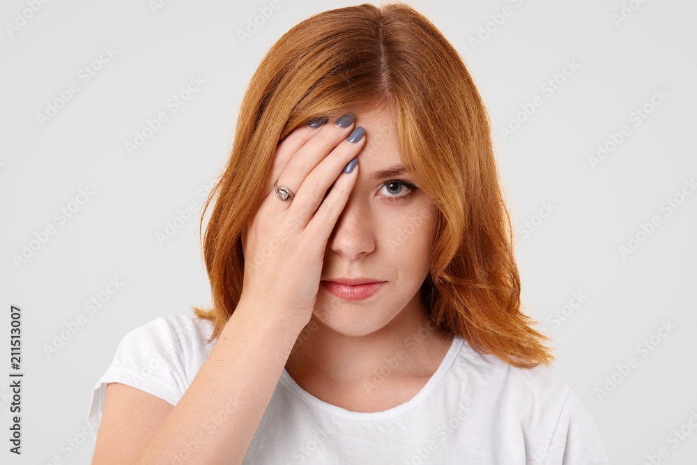 Angry sad female covers half of face after quarrel with boyfriend, being in low spirit, has freckled skin, looks with unhappy expression at camera, isolated over white wall. Facial expressions concept Angry sad female covers half of face after quarrel with boyfriend, being in low spirit, has freckled skin, looks with unhappy expression at camera, isolated over white wall. Facial expressions concept