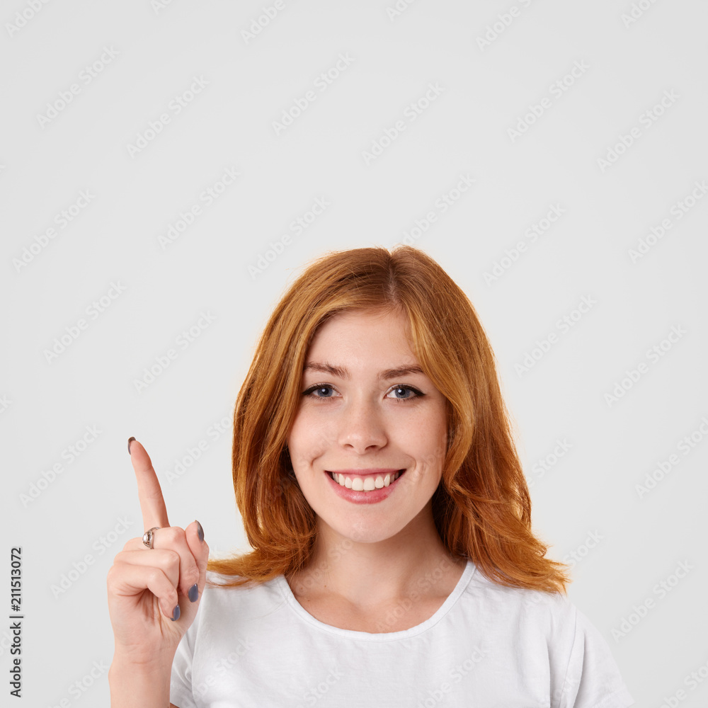 Positive lovely female with shining smile and red hair, points upwards with index finger, glad to present something, dressed in casual white t shirt, poses indoor alone. People and advertisement Positive lovely female with shining smile and red hair, points upwards with index finger, glad to present something, dressed in casual white t shirt, poses indoor alone. People and advertisement