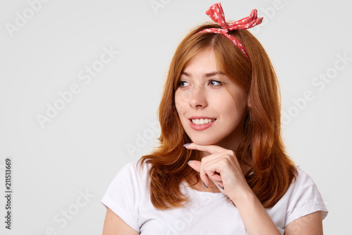 Happy Caucasian female with positive smile, holds chin and looks gladfully aside, wears stylish headband and white t shirt, poses against studio wall with copy space for your advertisement or text