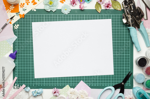 The cutting mat is surrounded by paper flowers, paper, tools and scrapbooking materials. Scrapbooking, top view, white blank sheet in the center