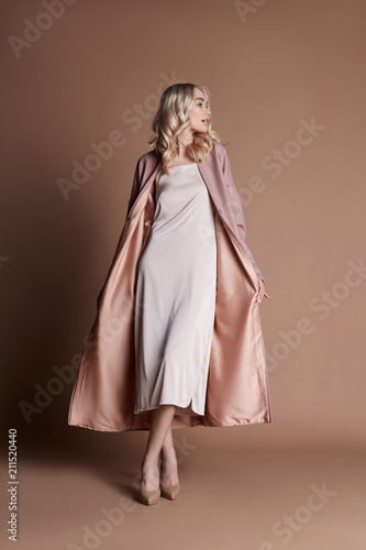 Beautiful blonde woman posing in a pink coat on a beige background. Fashion show clothing, woman with perfect figure, long hair. Trendy fashionable autumn coat on the girl's body