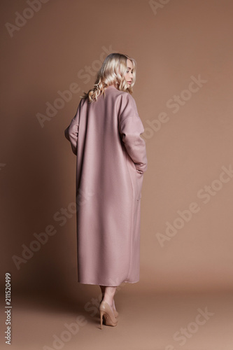 Beautiful blonde woman posing in a pink coat on a beige background. Fashion show clothing, woman with perfect figure, long hair. Trendy fashionable autumn coat on the girl's body