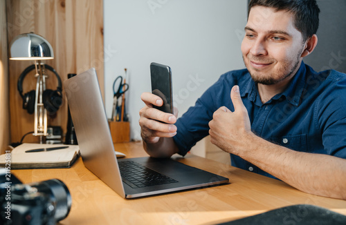 Young man in a blue shirt near a laptop on a wooden desk with a phone in his hand in the home office on a video link shows a gesture. Freelancer or photographer.