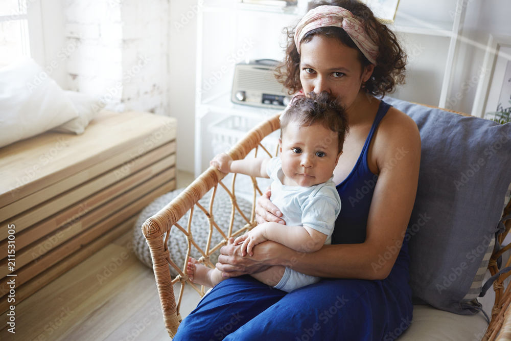 Indoor shot of charming young Latin mother wearing elegant blue dress  enjoying leisure time with toddler