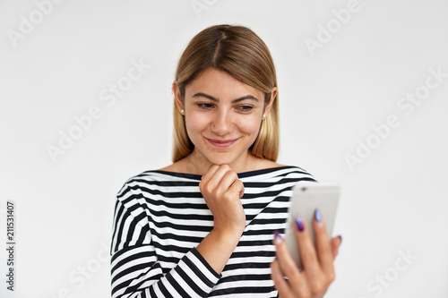Attractive young European female in striped top taking selfie using smart phone. Pretty girl speaking to friend via online video chat on electronic device. People, technology and communication