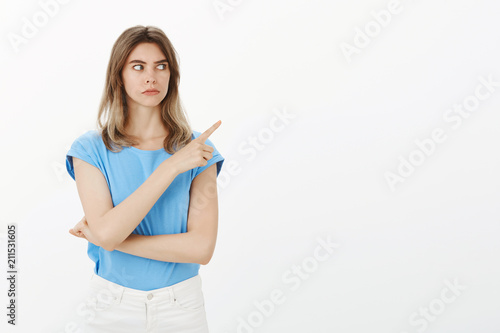Studio shot of intense worried young woman  staring and pointing right with index finger  seeing something weird and strange  being unsure what happening and asking question over grey wall