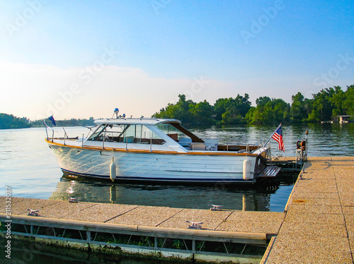A vintage wooden sea skiff boat with an American flag on the back moored at a dock in a lake with reflection an bouys and trees on the opposite bank