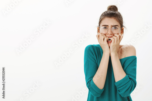 Studio shot of shocked and scared young woman in transparent glasses and loose sweater, frowning, biting fingernails, feeling nervous and anxious while being afraid facing destiny over grey wall