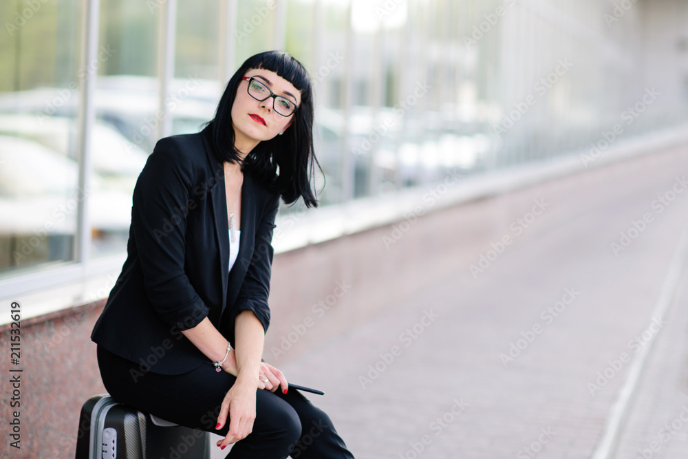 Young pretty girl dressed in a business suit and in the glasses with a suitcase talking on the phone and photographed