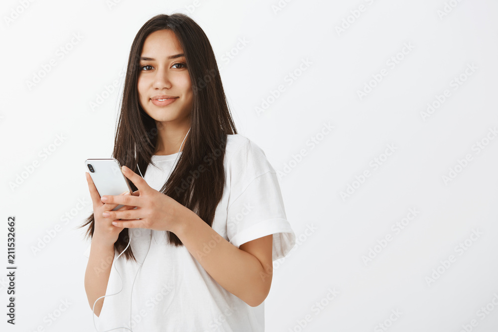 Waist-up shot of good-looking confident female coworker in white t-shirt, holding smartphone, listening music in earphones, smiling with tender and caring expression, enjoying nice songs Waist-up shot of good-looking confident female coworker in white t-shirt, holding smartphone, listening music in earphones, smiling with tender and caring expression, enjoying nice songs
