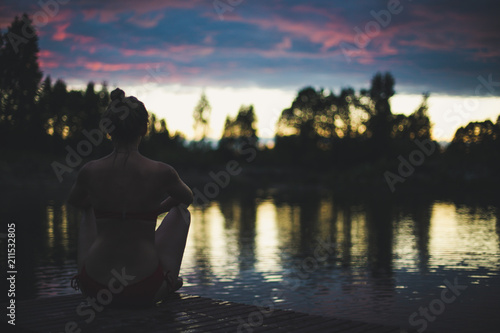 The woman is sitting on the footbridge at the lake in evening. Yoga concept. Vintage blur effect.