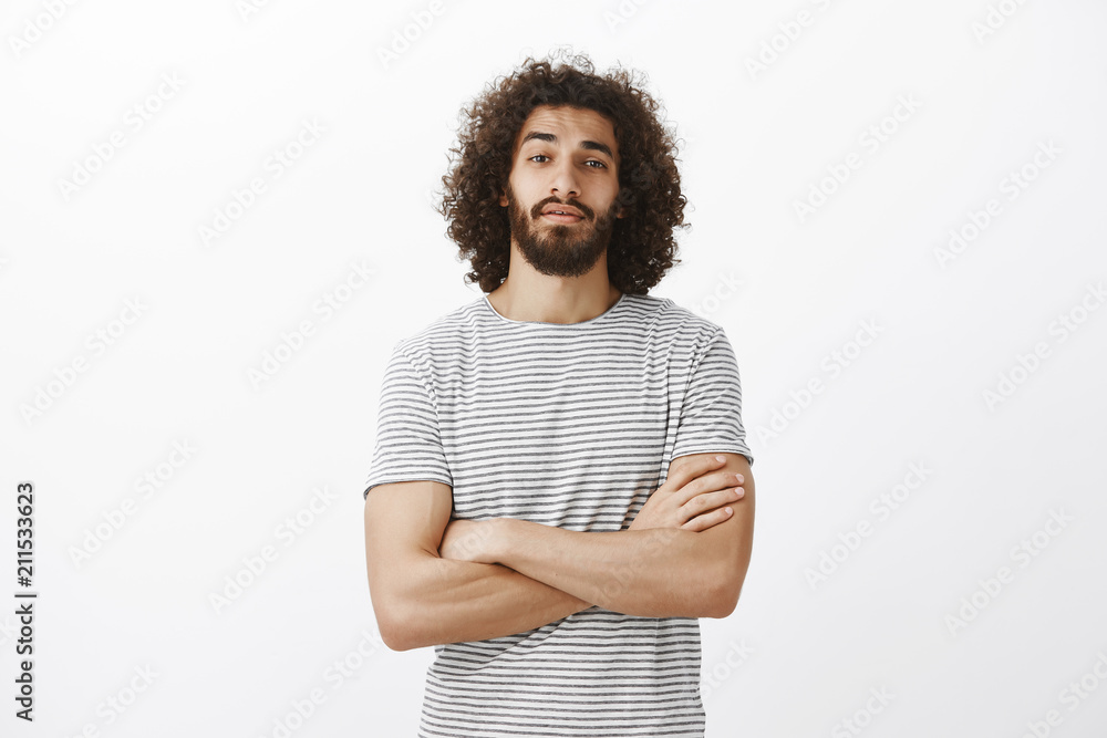 Indoor shot of popular handsome male model with beard and afro hairstyle, holding hands crossed on chest and looking with raised chin at camera, being confident and stubborn, standing over gray wall Indoor shot of popular handsome male model with beard and afro hairstyle, holding hands crossed on chest and looking with raised chin at camera, being confident and stubborn, standing over gray wall