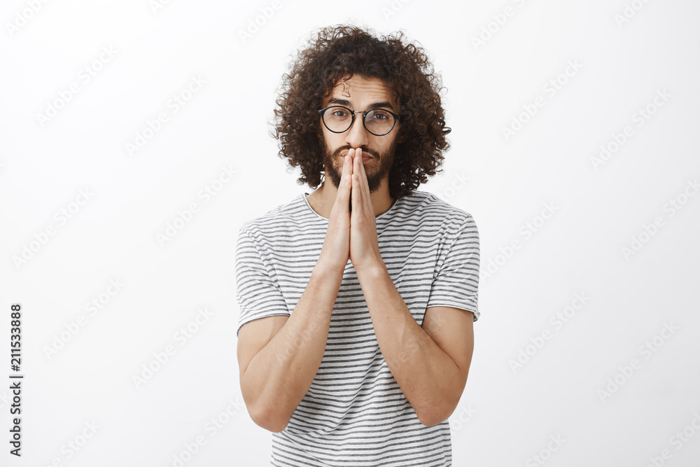 Confident good-looking hispanic guy with curly hair in trendy black glasses, holding hands in pray over mouth and nose and staring at camera, hoping for something and waiting calm for result