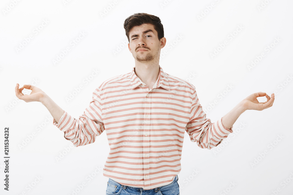 Fototapeta premium Studio shot of unfocused handsome male with moustache, spread hands in zen gesture and peeking with one eye, being calm and relaxed during meditation and yoga over gray background