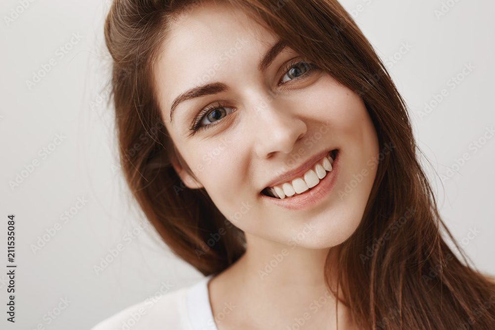Close-up shot of charmed good-looking caucasian girl tilting head from interest and curiosity, showing pleasure to talk with speaker, smiling broadly at camera, gazing with admiration over gray wall Close-up shot of charmed good-looking caucasian girl tilting head from interest and curiosity, showing pleasure to talk with speaker, smiling broadly at camera, gazing with admiration over gray wall