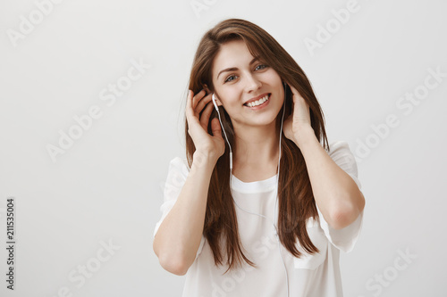 She loves music. Indoor shot of good-looking european student with brown hair wearing earphones and smiling broadly at camera, listening songs and enjoying good vibes over gray background