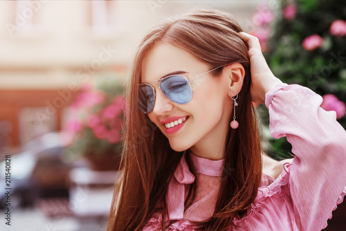 Outdoor close up portrait of young beautiful happy smiling woman wearing blue aviator sunglasses, trendy earrings, posing in street of european city. Copy, empty space for text