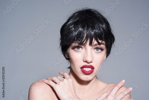 Beautiful woman with stylish make-up, big green eyes, short glossy black hair isolated on gray background. Naked female caressing her skin, embracing her shoulders, sensuality and tenderness concept
