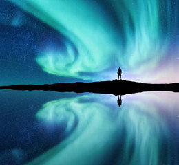 Gordijnen Koraalgroen Northern lights and silhouette of standing man in the hill in Norway. Aurora borealis and man. Stars and green polar lights. Night landscape with aurora, lake, sky reflection in water. Travel. Concept  © den-belitsky