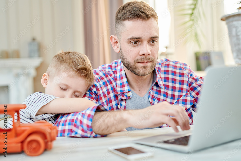 Warm toned portrait of cute little boy falling asleep hugging his dad waiting for him to finish working at laptop Warm toned portrait of cute little boy falling asleep hugging his dad waiting for him to finish working at laptop