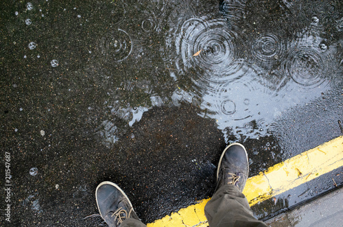 Rainy weather. Male legs in sneakers or boots walking through the rain puddle on the asphalt road, top view. Film grain photo.