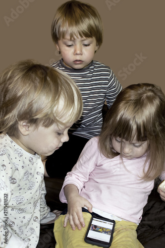 Little cute curious blonde triplets watch cartoons on smartphone indoors, half resaturated image isolated on dark beige photo