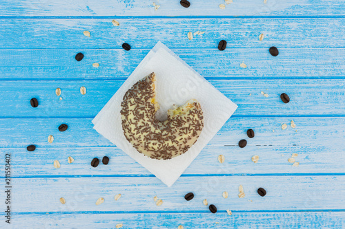 Bitten Tasty Sweet Donut On Paper On Scratched Blue White High Contrast Wood Background With Whole Grains Oat Meal And Coffee Beans On It