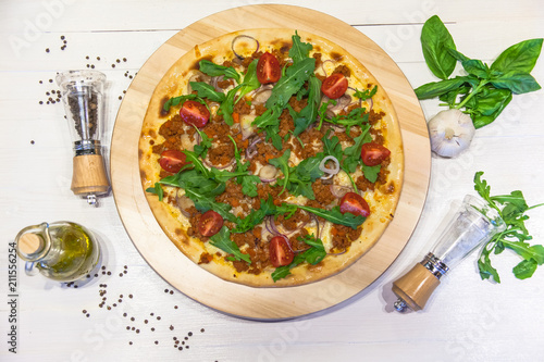 appetizing pizza with minced meat, small tomatoes, arugula spices and olive oil on a white table, flat lay