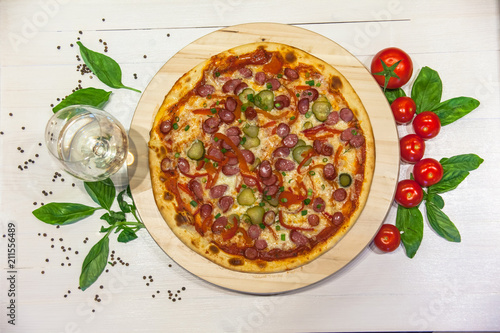 appetizing pizza with small sausages, basil, tomatoes and a glass of white wine on a white table, flat lay