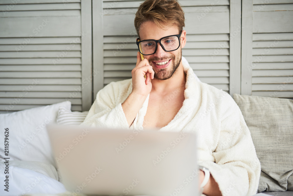 Waist up portrait of happy young male sitting on bed in bathrobe and holding laptop. He is communicating on mobile while looking at computer screen Waist up portrait of happy young male sitting on bed in bathrobe and holding laptop. He is communicating on mobile while looking at computer screen