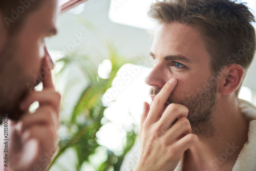 Side view profile of young man standing in front of mirror. He is looking precisely at face and touching skin under eyes with finger