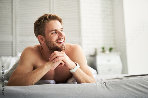 Perfect morning. Waist up portrait of happy male lying on belly and looking sideways. He is holding hands together and smiling admiring window view