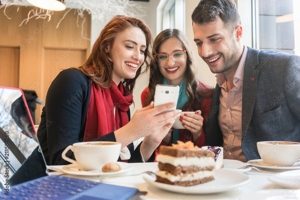 Three young colleagues or friends smiling while using a mobile phone for fun on social media during a coffee break in a trendy cafeteria