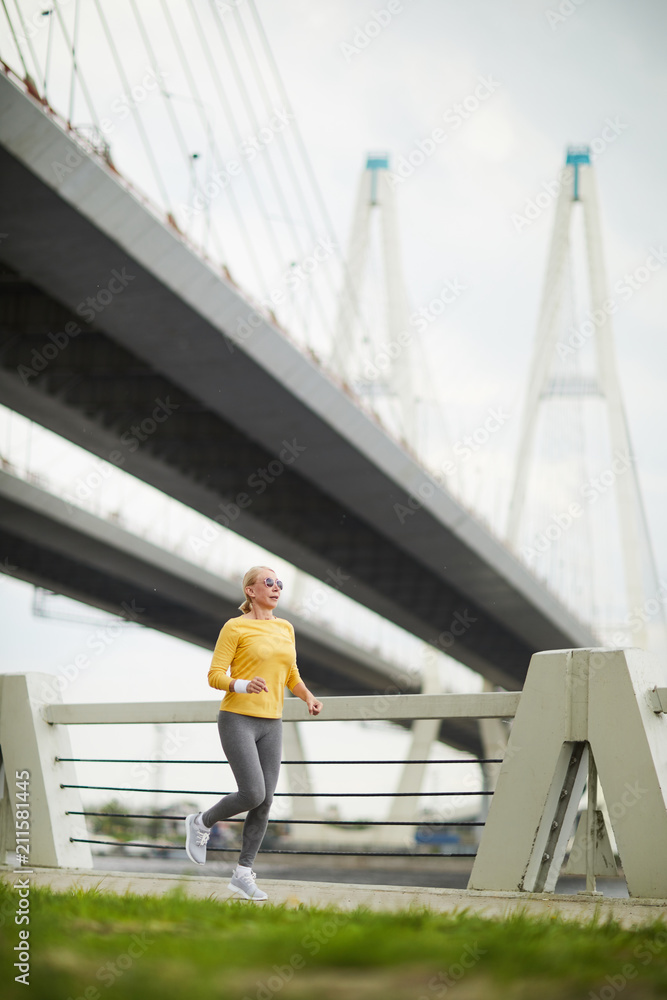 Active senior female wearing yellow pullover and grey leggins jogging along bridge outdoors