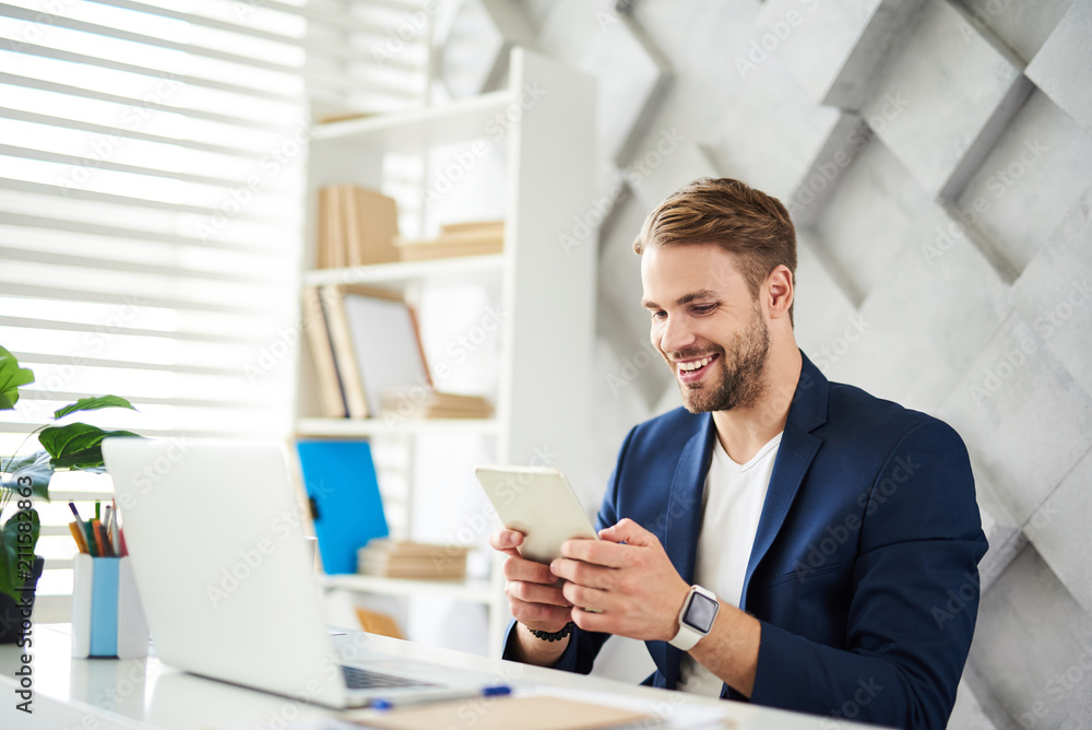 Side view of happy male sitting at table with computer on it. He is using portative device for fun whenever he has free times Side view of happy male sitting at table with computer on it. He is using portative device for fun whenever he has free times