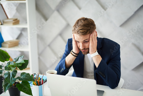 Oh my God. Confused businessman working on project at computer. He is holding head with hand and looking at screen in bewildering 