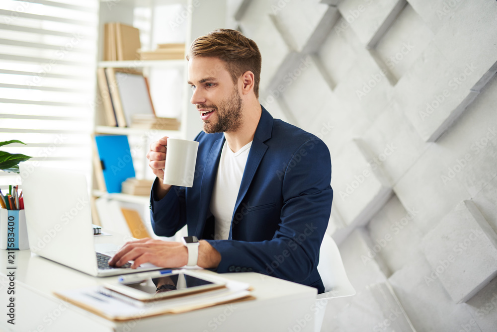 Side view of smiling male sitting at table with coffee mug in hands. He is looking at laptop enjoying work in business center Side view of smiling male sitting at table with coffee mug in hands. He is looking at laptop enjoying work in business center