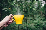 Hand holding hot yellow coffee cup with hand drawn smile face on cup at tropical nature forest,Leisure lifestyle.